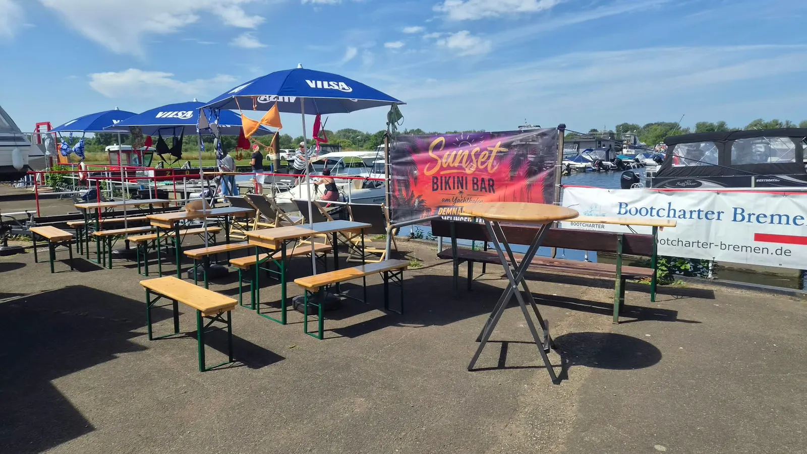 Biertischgarnituren und Sonnenschirme am Hafenbecken, dahinter das Banner der Sunset Bikini Bar
