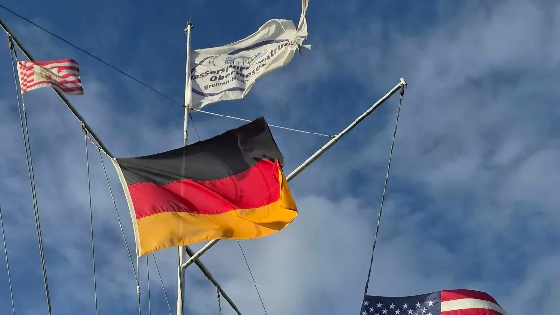 Am Flaggenmast wehen der Wimpel des Wassersport-Zentrums Oberweser, die Bremer Speckflagge und die Deutschlandflagge vor blauem Himmel
