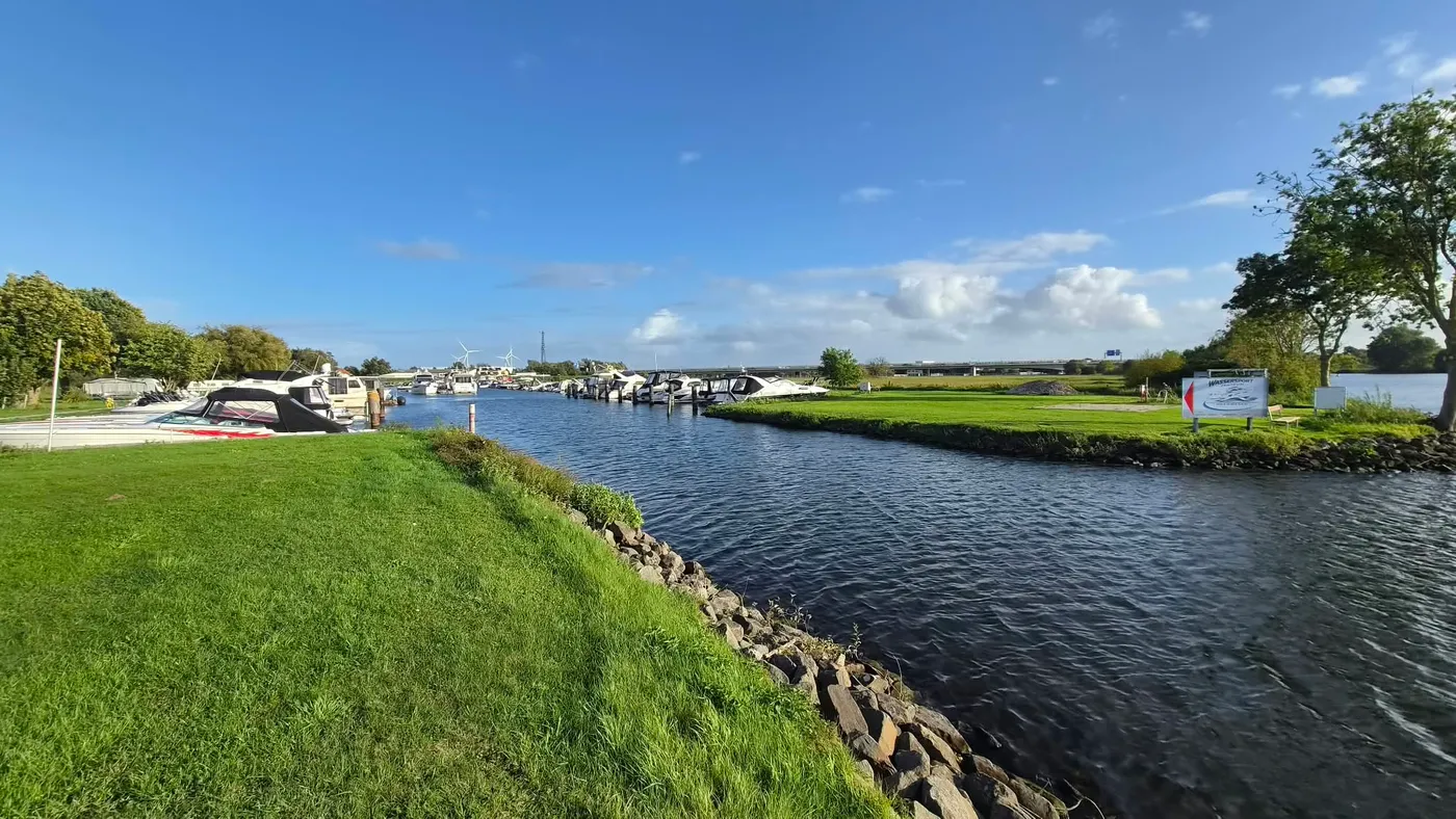 Blick durch die Hafeneinfahrt: links liegen Boote am Ufer, rechts die Wiese mit dem Hafenschild, dahinter die Weser