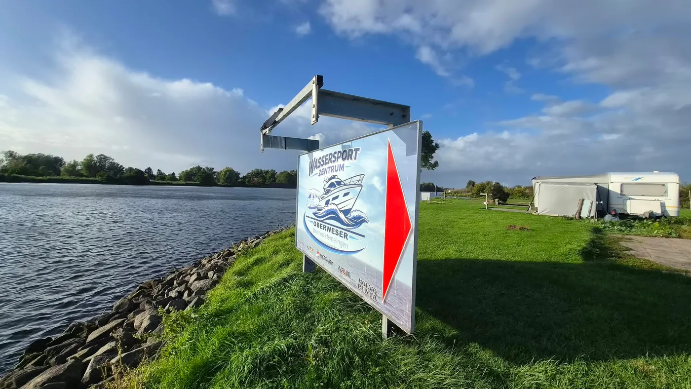 Das Schild „Wassersport-Zentrum Oberweser“ steht am Weserufer über der Steinschüttung, dahinter die Stellplatzwiese