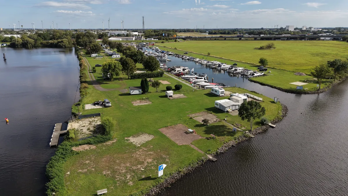 Luftaufnahme der Stellplatzwiese zwischen Weser und Hafenbecken: Wohnwagen und Wohnmobile stehen im Grünen zwischen Bäumen, dahinter die Boote am Steg