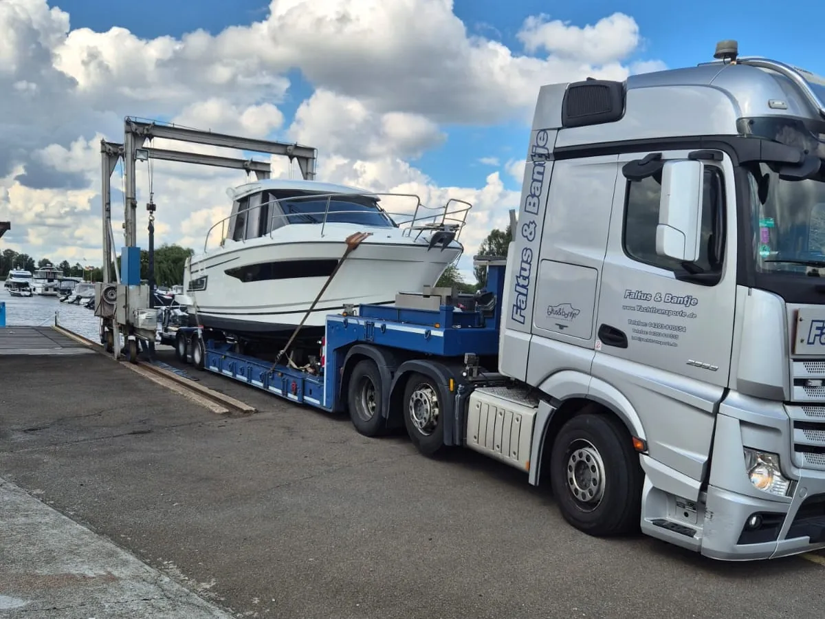 Eine weiße Motoryacht steht festgezurrt auf dem Tieflader eines Yachttransporters, dahinter der Hafenlift und Boote im Hafenbecken